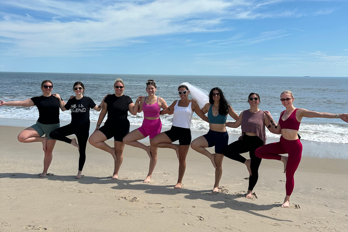 Group of women doing yoga tree pose on sandy beach with ocean and blue sky